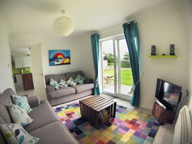 A living room with two sofas and colorful cushions a wooden coffee table a multicolored rug a TV with speakers and a sliding glass door with blue curtains at Driftwood in Bude