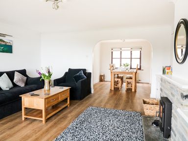A living room with black sofas and a wooden coffee table next to a fireplace and a dining area with a wooden table and chairs at Seal Cottage in Mortehoe