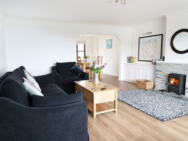 A living room with a black sofa and armchair a wooden coffee table and a fireplace a dining area visible through an archway at Seal Cottage in Mortehoe