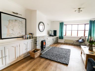 A living room with a fireplace television chair and a large window with blue curtains at Seal Cottage in Mortehoe
