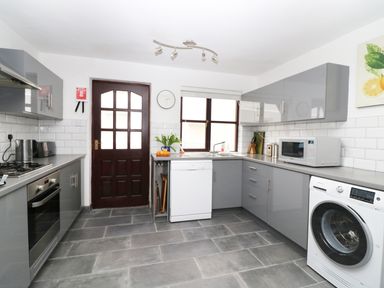 A kitchen with grey cabinets a wooden door a dishwasher a washing machine and a microwave at Seal Cottage in Mortehoe