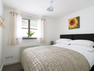 A bedroom with a double bed with white pillows and beige bedspread near a window with curtains and a sunflower painting on the wall at Seal Cottage in Mortehoe