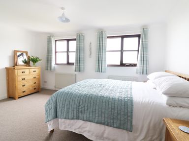 A bedroom with a double bed covered with a blue bedspread two windows with blue plaid curtains and a wooden dresser with a mirror and flowers at Seal Cottage in Mortehoe