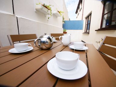 An outdoor patio with a wooden table set with white cups saucers a silver teapot and a white jug at Seal Cottage in Mortehoe