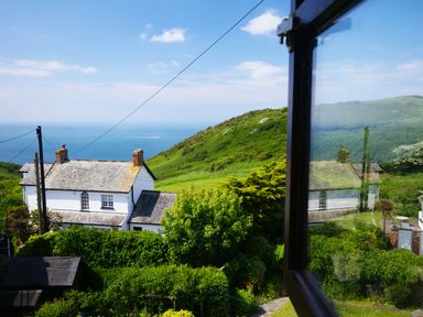 View of a white house with chimneys surrounded by green vegetation and hills with the sea in the background at Seal Cottage in Mortehoe