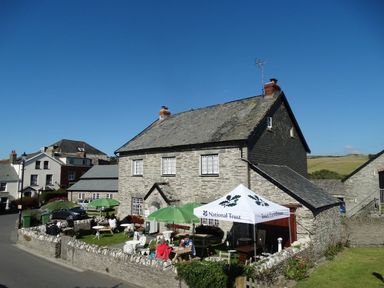 An outdoor seating area with tables umbrellas and a National Trust tent outside a stone farmhouse at Seal Cottage in Mortehoe