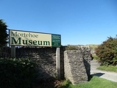 An outdoor sign for Mortehoe Museum next to a stone wall and bushes at Mortehoe