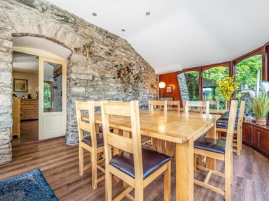 A dining room with a wooden table and chairs near stone walls and a large window with plants at Eden Mill Millers Beck in Kendal