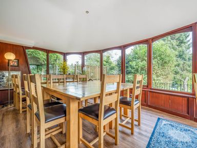 A dining room with a large wooden table and chairs surrounded by windows overlooking trees at Eden Mill Millers Beck in Kendal