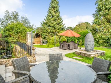 An outdoor garden area with round glass tables, wicker chairs, an orange umbrella, stone path, greenery, and a water feature at Eden Mill Millers Beck in Kendal
