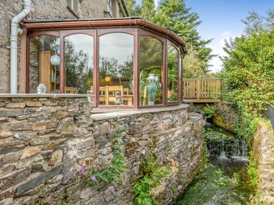 A stone wall with a glass conservatory above overlooking a narrow stream with vegetation at Eden Mill Millers Beck in Kendal