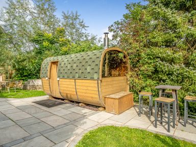 An outdoor area with a wooden barrel-shaped structure surrounded by trees and a patio with tall stools and tables at Eden Mill Millers Beck in Kendal