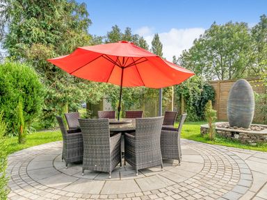 A circular outdoor patio with wicker chairs and a table under a red umbrella near a garden and stone water feature at Eden Mill Millers Beck Kendal