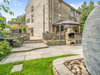 An outdoor patio area with stone paving a stone barbecue and seating beside a stone building with a conservatory at Eden Mill Millers Beck in Kendal