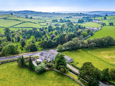 An aerial view of a rural stone house with surrounding trees fields and distant hills at Eden Mill Millers Beck in Kendal