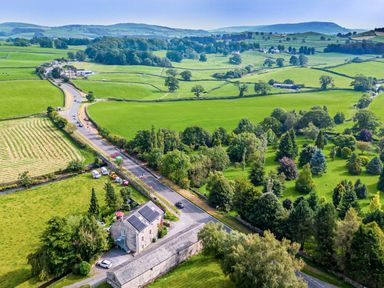 A road passing through green fields and trees with houses and hills in the background at Eden Mill Millers Beck in Kendal