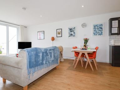A living area with a beige sofa covered by a blue throw facing a television and a dining table with four orange chairs at Buena Vista in Newquay