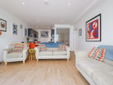 A living room with two sofas and an armchair with patterned cushions and blue throws and a kitchen area with black cabinets and a dining table with orange chairs at Buena Vista in Newquay