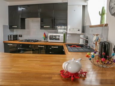 A kitchen with black cabinets a wooden countertop a white teapot a colorful fruit bowl a microwave and a sink at The Compass in Swanage