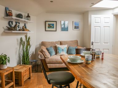 A small living area with a sofa pillows dining table with dishes and mugs wooden stools and shelves with decorations at The Compass in Swanage