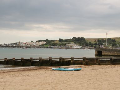 A sandy beach with a paddleboard on it by a wooden groyne and a pier with boats in the water and houses on a hill at The Compass in Swanage