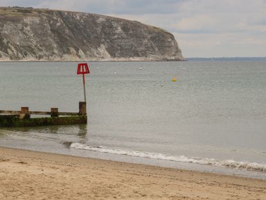 A sandy beach with a seawall and a red navigational marker in the sea with cliffs in the background at The Compass in Swanage