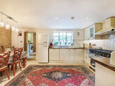 A kitchen with white cabinets a white refrigerator a dining table with red chairs and a red patterned rug on the floor at Puffitts Cottage in Bourton-on-the-Water