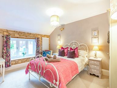 A bedroom with a white metal bed frame pink and purple pillows a pink bed cover two bedside tables with lamps floral curtains and a window at Puffitts Cottage in Bourton-on-the-Water