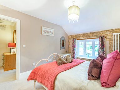 A bedroom with a bed covered with pillows and a red blanket near a window with floral curtains at Puffitts Cottage in Bourton-on-the-Water
