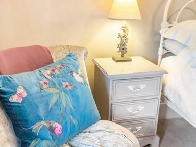 A bedroom corner with a padded chair holding a blue decorative cushion and a white bedside table with a lamp at Puffitts Cottage in Bourton-on-the-Water
