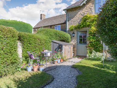A garden with a gravel path leading to a door of a stone house with potted plants and a small table with chairs at Puffitts Cottage in Bourton-on-the-Water