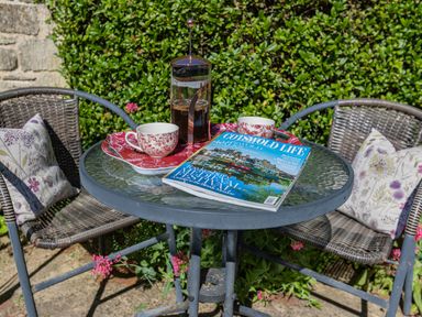 A glass outdoor table with a French press coffee maker two cups and a magazine between two wicker chairs with floral cushions at Puffitts Cottage Bourton-on-the-Water