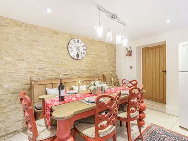 A dining room with a wooden table set with plates candles and a bottle of wine four red chairs a wooden bench stone wall with clock and wooden door in the background at Puffitts Cottage in Bourton-on-the-Water