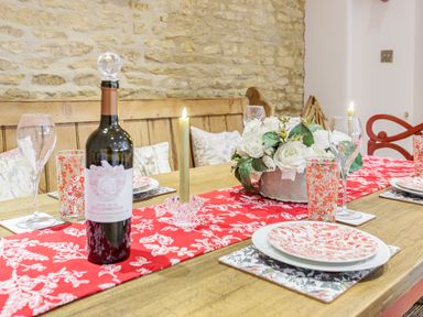 A dining table set with a red and white table runner wine bottle candles floral centerpiece and plates at Puffitts Cottage in Bourton-on-the-Water