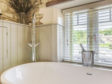 A bathtub with a chrome faucet near a window with blinds and a silver ice bucket with a bottle and two glasses at Puffitts Cottage in Bourton-on-the-Water