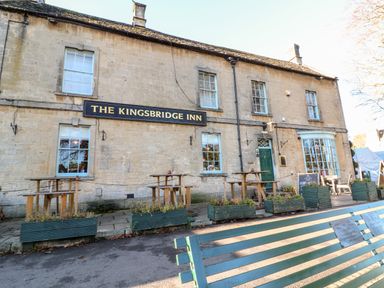The exterior of The Kingsbridge Inn with wooden tables and planters outside and a green bench in front at Puffitts Cottage in Bourton-on-the-Water