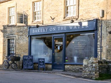 The front of Bakery On The Water with stone walls a bicycle and two chalkboard signs outside at Puffitts Cottage in Bourton-on-the-Water