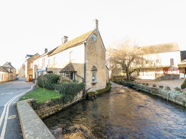 A stone building beside a flowing river with a paved road and other buildings in the background at Puffitts Cottage in Bourton-on-the-Water