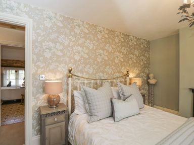 A bedroom with a bed and pillows two bedside tables with lamps floral wallpaper and a bust on a pedestal at Puffitts Cottage in Bourton-on-the-Water