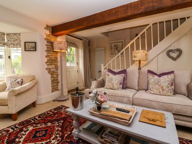 A living room with a beige sofa and armchair a patterned rug and a coffee table with flowers and a wine bucket at Puffitts Cottage in Bourton-on-the-Water