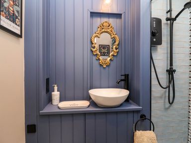 A bathroom sink area with a round white basin black faucet gold mirror and blue panel wall at Puffitts Cottage in Bourton-on-the-Water
