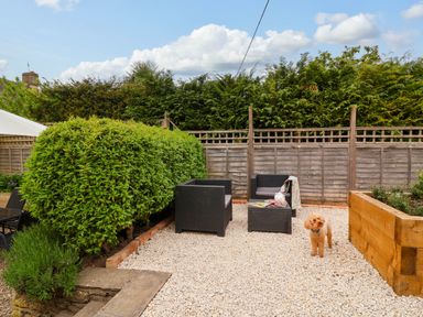 A backyard seating area with black wicker chairs and table on gravel near wooden planter and green bushes at Puffitts Cottage in Bourton-on-the-Water
