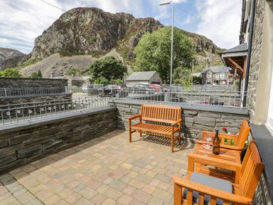 An outdoor patio area with wooden benches a table with a bottle and wine glasses stone walls and a mountain in the background at Garreg Ddu in Blaenau Ffestiniog
