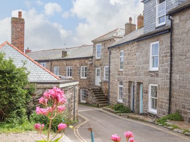 A narrow street with stone houses and chimneys with pink flowers in the foreground at Mermaid Cottage in Mousehole