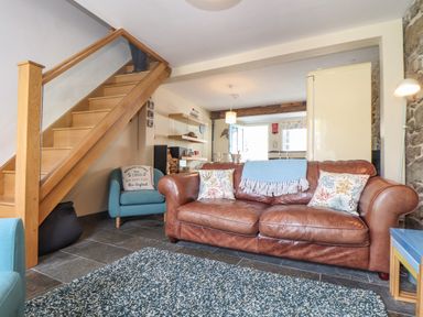 A living room with a brown leather sofa blue armchairs a wooden staircase and stone wall at Mermaid Cottage in Mousehole