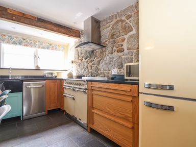 A kitchen with wooden cabinets a stone wall a cream refrigerator a stove a stainless steel dishwasher and a window with a patterned valance at Mermaid Cottage in Mousehole