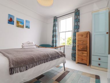 A bedroom with a bed and folded towels on it a chest of drawers a cupboard a window with patterned curtains and paintings on the wall at Mermaid Cottage in Mousehole