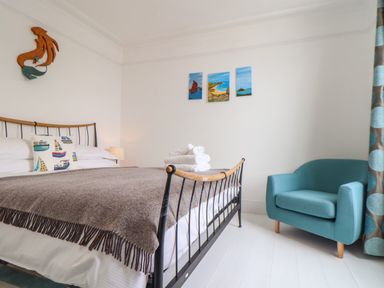 A bedroom with a bed with a brown throw and boat themed cushion a blue armchair and paintings on the wall at Mermaid Cottage in Mousehole