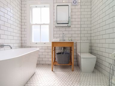 A bathroom with a bathtub a wooden sink stand with a basket a toilet and a window at Mermaid Cottage in Mousehole
