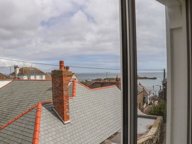A view of rooftops with chimneys and the sea in the background seen through a window at Mermaid Cottage in Mousehole